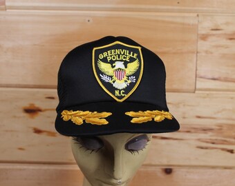 May include: Black mesh trucker hat with a gold embroidered patch that says "Greenville Police" and features an eagle with an American flag. The patch also has the letters "N.C."