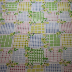 May include: A white fabric with a patchwork design featuring squares of blue, green, yellow, and pink gingham patterns. Each square is outlined in a thin brown line and decorated with small floral clusters.