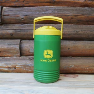 May include: A green insulated beverage cooler with a yellow lid and a yellow swing handle. The front features the yellow John Deere logo and text. The cylindrical body has horizontal ridges at the base and a smooth upper section for branding.