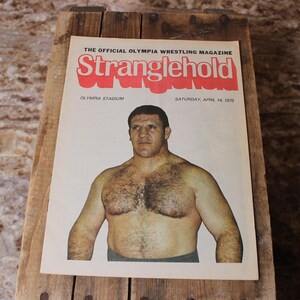 Pode incluir: Capa de revista de luta livre vintage com um homem sem camisa. O título da revista é "Stranglehold" em letras vermelhas. A capa também inclui o texto "Olympia Stadium" e "Saturday, April 14, 1973". A revista está sobre uma superfície de madeira.