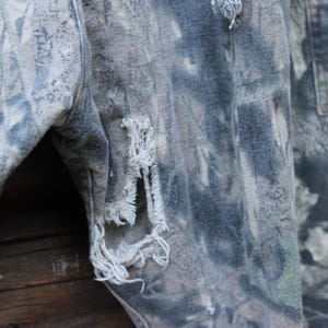 May include: Close-up of camouflage fabric with distressed details. The fabric has green, grey, and brown hues. Frayed edges and tears are visible, giving a worn, vintage look. The texture suggests a durable material, possibly denim or canvas.