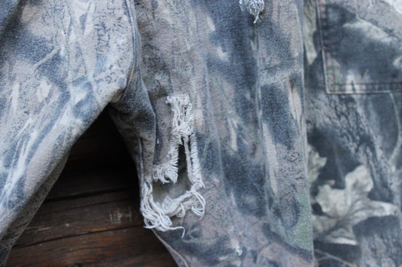May include: Close-up of camouflage fabric with distressed details. The fabric has green, grey, and brown hues. Frayed edges and tears are visible, giving a worn, vintage look. The texture suggests a durable material, possibly denim or canvas.