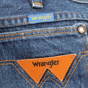 May include: Close-up of a pair of blue denim jeans with a brown leather Wrangler patch on the back pocket. The patch has the word "Wrangler" in white lettering.