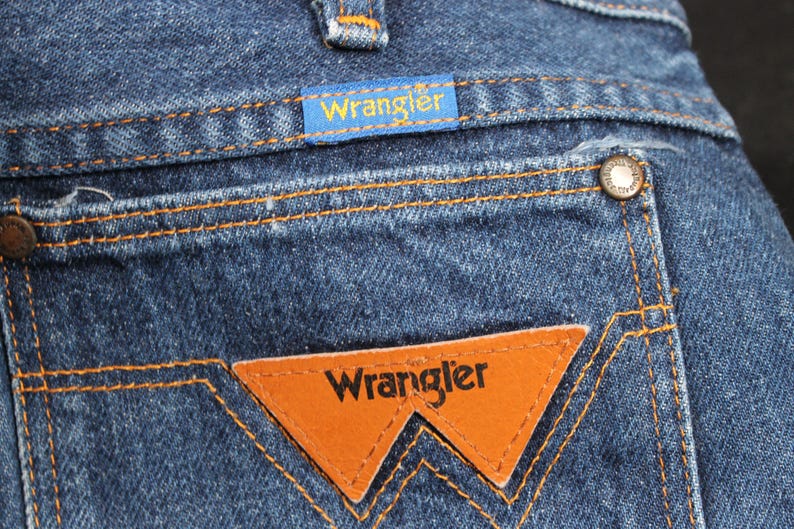 May include: Close-up of a pair of blue denim jeans with a brown leather Wrangler patch on the back pocket. The patch has the word "Wrangler" in white lettering.