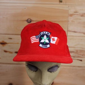 Könnte beinhalten: Rote Cord-Baseballkappe mit einem gestickten Aufnäher mit den Buchstaben "I.B.E.W." und einer weißen Blume mit grünen Blättern, umgeben von einer kleinen amerikanischen Flagge und einer kanadischen Flagge. Der Aufnäher enthält auch das Jahr "1789".