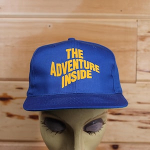 Könnte beinhalten: Eine blaue Baseballkappe mit einem gelben gestickten Design, das "The Adventure Inside" lautet.