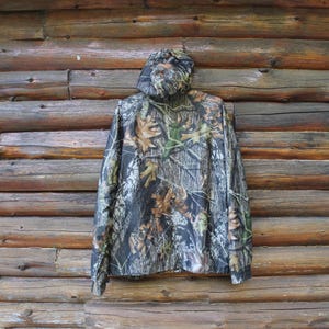 May include: A camouflage hooded jacket with a woodland pattern of brown, green, and black. The jacket is hanging against a rustic wooden log wall. The jacket has a full zipper and a hood.