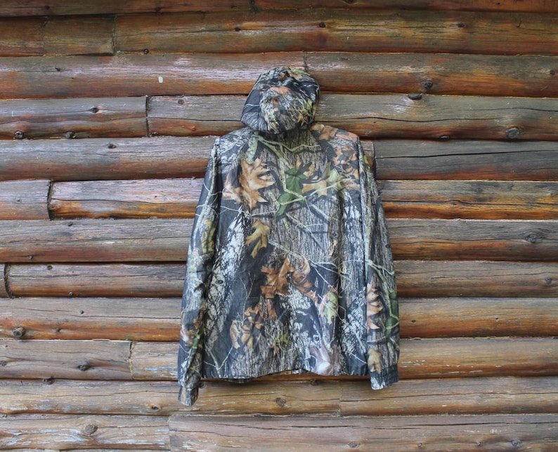 May include: A camouflage hooded jacket with a woodland pattern of brown, green, and black. The jacket is hanging against a rustic wooden log wall. The jacket has a full zipper and a hood.