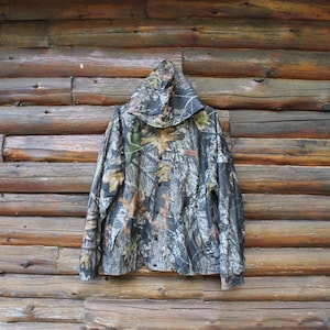 May include: A hooded camouflage jacket with a button-up front, featuring a pattern of brown, green, and gray tones. The jacket is hanging against a rustic wooden log wall, showcasing its outdoor-oriented design.