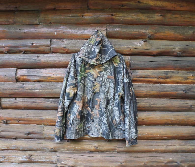 May include: A hooded camouflage jacket with a button-up front, featuring a pattern of brown, green, and gray tones. The jacket is hanging against a rustic wooden log wall, showcasing its outdoor-oriented design.