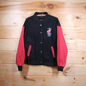 K&ouml;nnte beinhalten: Schwarze und rote Baseballjacke mit einer Comicfigur auf der Vorderseite. Die Figur tr&auml;gt einen roten Anzug und eine blaue M&uuml;tze. Der Text "La Bamba" befindet sich &uuml;ber der Figur und "Professional" und "GOURMET" unter der Figur.