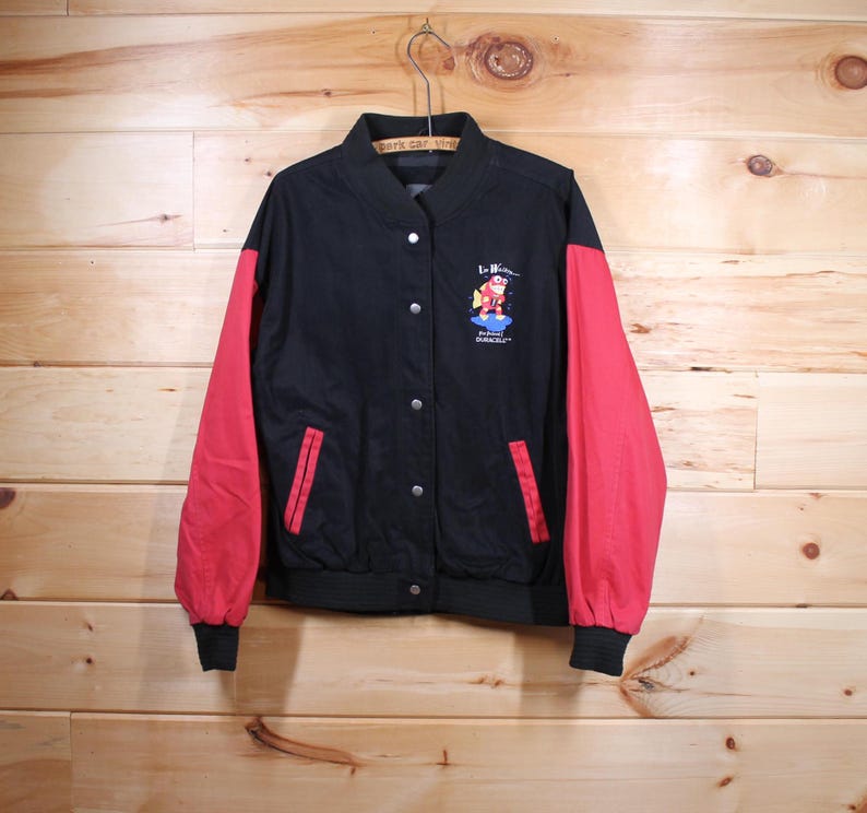 K&ouml;nnte beinhalten: Schwarze und rote Baseballjacke mit einer Comicfigur auf der Vorderseite. Die Figur tr&auml;gt einen roten Anzug und eine blaue M&uuml;tze. Der Text "La Bamba" befindet sich &uuml;ber der Figur und "Professional" und "GOURMET" unter der Figur.