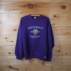 Puede incluir: Una sudadera morada con cuello redondo con el texto "UNIVERSITY OF TORONTO" en blanco, con un emblema circular. La sudadera está colgada de una percha de madera sobre un fondo de madera. El texto "established in 1827" está debajo.