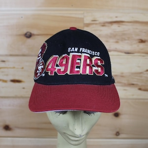 May include: Black baseball cap with a red peak. The cap features the text "SAN FRANCISCO 49ERS" in white and red embroidery. The hat is displayed on a mannequin head against a wooden background.