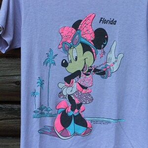 May include: A light purple t-shirt featuring a graphic of Minnie Mouse wearing pink and blue sunglasses, a bow, and a patterned outfit. The design includes the text Florida, palm trees, and star-shaped earrings in a vibrant neon color palette.