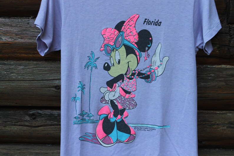 May include: A light purple t-shirt featuring a graphic of Minnie Mouse wearing pink and blue sunglasses, a bow, and a patterned outfit. The design includes the text Florida, palm trees, and star-shaped earrings in a vibrant neon color palette.