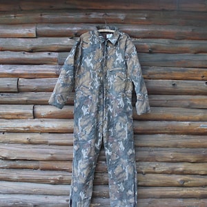 Puede incluir: Mono con estampado de camuflaje con cuello y mangas largas. La prenda de una pieza presenta cierre de cremallera y m&uacute;ltiples bolsillos. Los colores incluyen tonos de marr&oacute;n, verde y gris, creando un dise&ntilde;o de camuflaje de bosque. El mono est&aacute; colgado de una percha de madera.