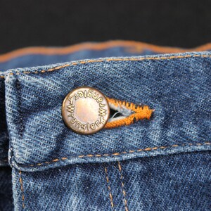 May include: Close-up of a blue denim jean waistband with a copper-colored button that reads "Wrangler".