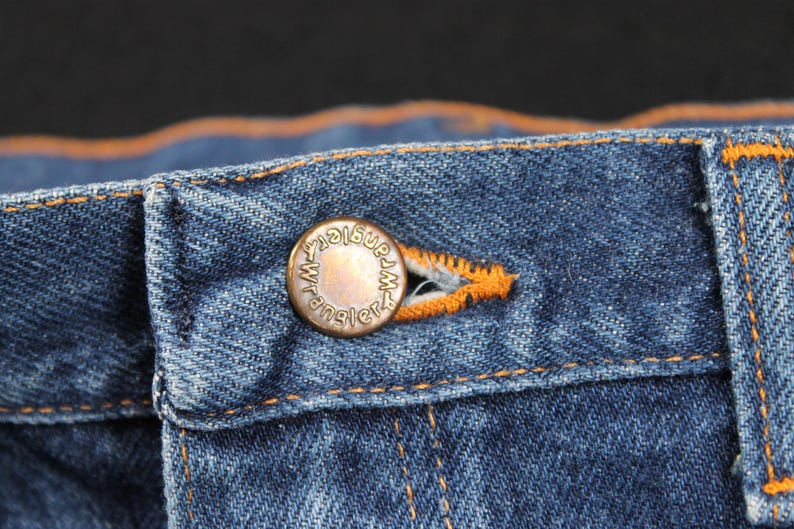 May include: Close-up of a blue denim jean waistband with a copper-colored button that reads "Wrangler".