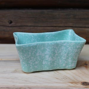 May include: A small, rectangular, turquoise ceramic planter with a speckled white pattern. The planter has a unique, wavy rim and sits on a wooden surface. The background is a rustic wooden wall.