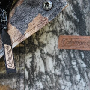 May include: Close-up of a camouflage jacket featuring a black zipper and a brown leather patch with the "Coleman" logo. The jacket has a button closure and a black tag with the "Coleman" logo.