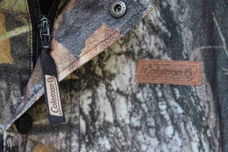 May include: Close-up of a camouflage jacket featuring a black zipper and a brown leather patch with the "Coleman" logo. The jacket has a button closure and a black tag with the "Coleman" logo.