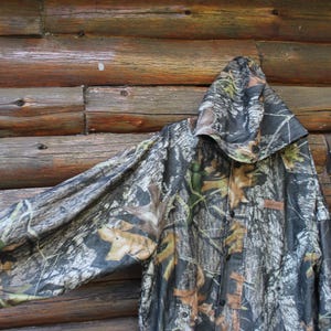 May include: Camouflage hooded jacket with a button-up front. The jacket features a woodland camouflage pattern in shades of brown, green, and gray. The jacket is hanging against a rustic wooden background.