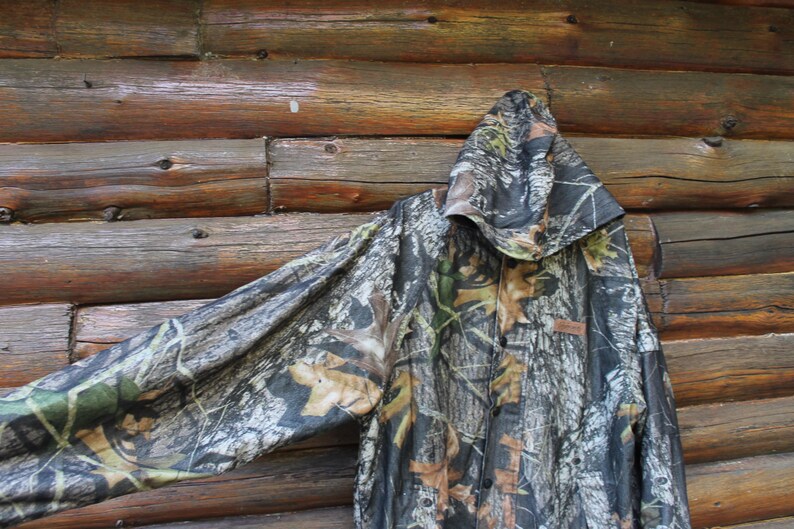 May include: Camouflage hooded jacket with a button-up front. The jacket features a woodland camouflage pattern in shades of brown, green, and gray. The jacket is hanging against a rustic wooden background.