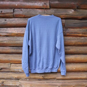 Puede incluir: Una sudadera azul claro cuelga de una percha de madera sobre un fondo de troncos de madera r&uacute;sticos. La parte superior de manga larga tiene cuello redondo y pu&ntilde;os y dobladillo acanalados. La tela parece suave y ligeramente arrugada.