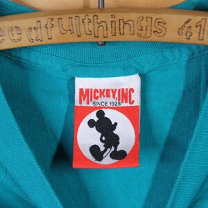 May include: A turquoise blue t-shirt with a red and white Mickey Mouse Inc. label. The label has the text "Mickey, Inc. Since 1928" and a black silhouette of Mickey Mouse.