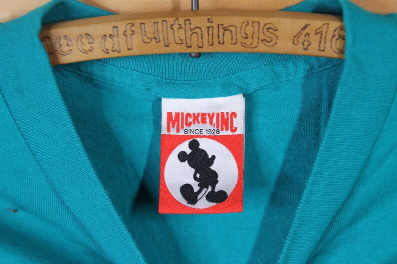 May include: A turquoise blue t-shirt with a red and white Mickey Mouse Inc. label. The label has the text "Mickey, Inc. Since 1928" and a black silhouette of Mickey Mouse.