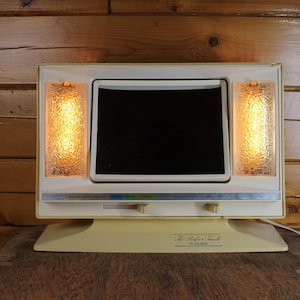Puede incluir: Un televisor vintage de color crema con una pantalla negra y dos paneles de vidrio texturizado que se iluminan con una luz amarilla cálida. La base tiene el texto "The Perfect Touch" y un número de modelo.
