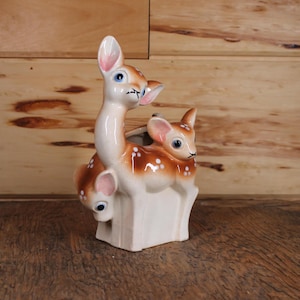 Puede incluir: Un macetero de cerámica con forma de ciervo madre y cervatillo. Los ciervos son marrones y blancos con manchas. El macetero es de color marrón claro.