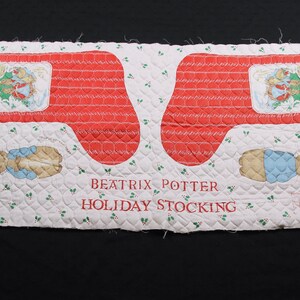 Op de afbeelding: Een wit stoffen paneel met een rood en wit gestikt patroon. Het paneel heeft twee rode kousvormen met een Peter Rabbit-illustratie en de tekst "Happy Holidays". Het paneel bevat ook de tekst "Beatrix Potter Holiday Stocking".