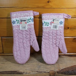 Peut inclure: Deux maniques de four matelassées rose clair. Chaque manique a une bande blanche à motifs floraux et de feuilles en haut, avec une boucle pour suspendre. Elles sont présentées sur une surface en bois.