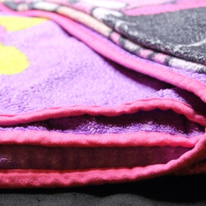 Pode incluir: Uma manta de l&atilde; polar roxa e rosa com um desenho de flor amarela. A manta est&aacute; dobrada ao meio e as bordas s&atilde;o vis&iacute;veis.