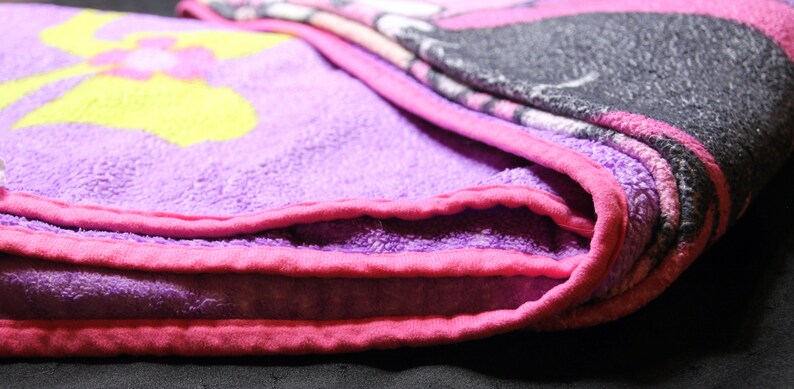 Pode incluir: Uma manta de l&atilde; polar roxa e rosa com um desenho de flor amarela. A manta est&aacute; dobrada ao meio e as bordas s&atilde;o vis&iacute;veis.