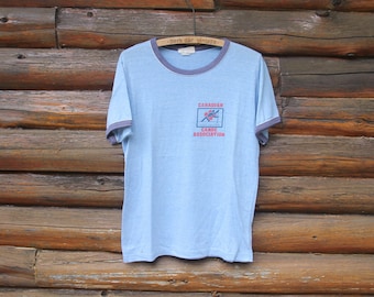 Könnte beinhalten: Ein hellblaues T-Shirt mit einem Ringkragen in Grau und Weiß. Das Shirt zeigt ein Vintage-Logo der Canadian Canoe Association mit einer roten und blauen Illustration eines Kanus und Paddlern.