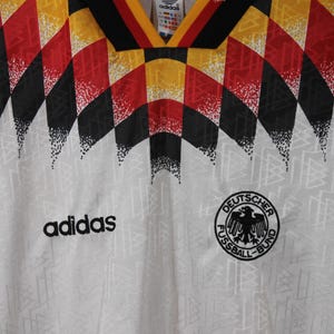 May include: White soccer jersey with a black and white geometric pattern, red and yellow accents, and the Adidas logo. The jersey also features the German national team logo, "Deutscher Fussball-Bund", with a black eagle.