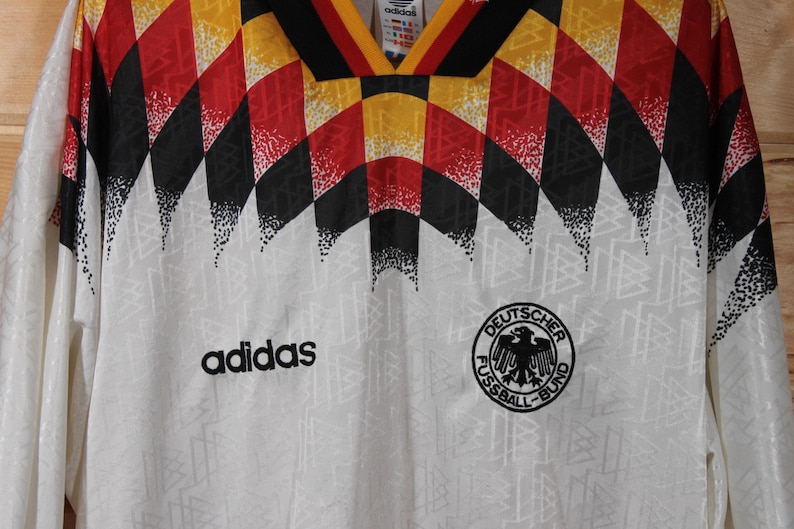 May include: White soccer jersey with a black and white geometric pattern, red and yellow accents, and the Adidas logo. The jersey also features the German national team logo, "Deutscher Fussball-Bund", with a black eagle.