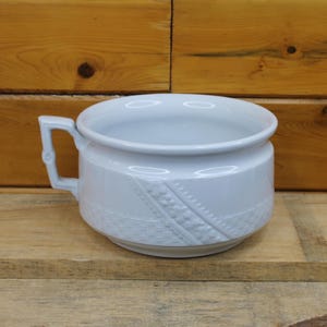 May include: A white ceramic chamber pot featuring a single angular handle and a decorative embossed band around the lower exterior. The vessel has a wide, open top and a smooth, glossy finish, showcasing a clean and functional design.