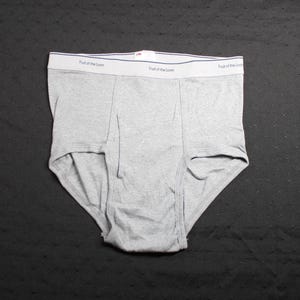 Peut inclure: Slip gris avec une ceinture blanche portant l'inscription "Fruit of the Loom" en noir. Le sous-vêtement est fait d'une matière douce et est posé à plat sur une surface sombre. Le slip a un design classique.