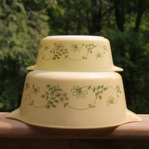 May include: A set of two vintage Pyrex mixing bowls with a floral design in green and white. The bowls are stacked on top of each other.