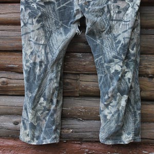 May include: Camouflage trousers in grey, brown, and green hues, with a tear near the crotch. The trousers are displayed against a weathered wooden backdrop, suitable for outdoor activities.