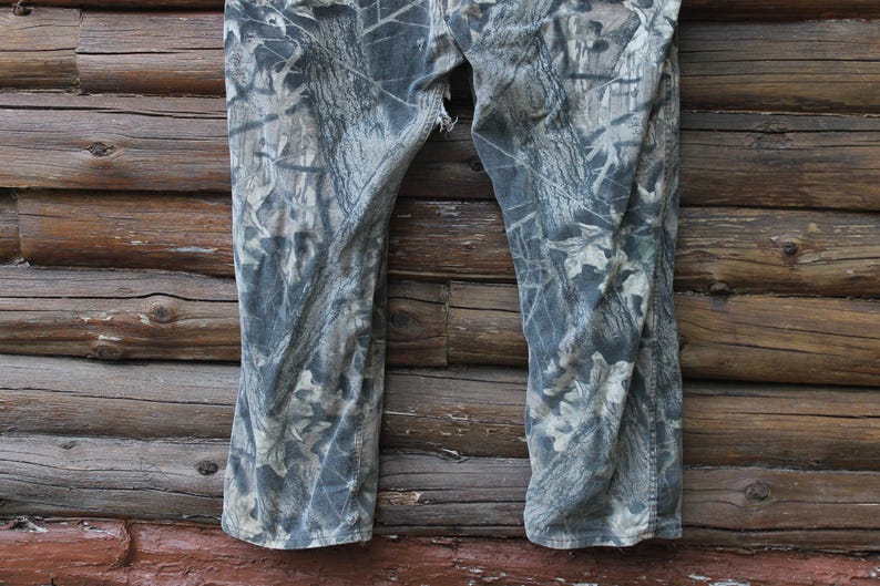 May include: Camouflage trousers in grey, brown, and green hues, with a tear near the crotch. The trousers are displayed against a weathered wooden backdrop, suitable for outdoor activities.