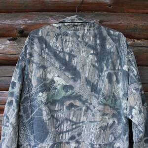May include: A camouflage jacket hangs on a hanger against a wooden background. The jacket features a pattern of greens, browns, and greys, typical of hunting apparel. The jacket has a collar and buttoned front.