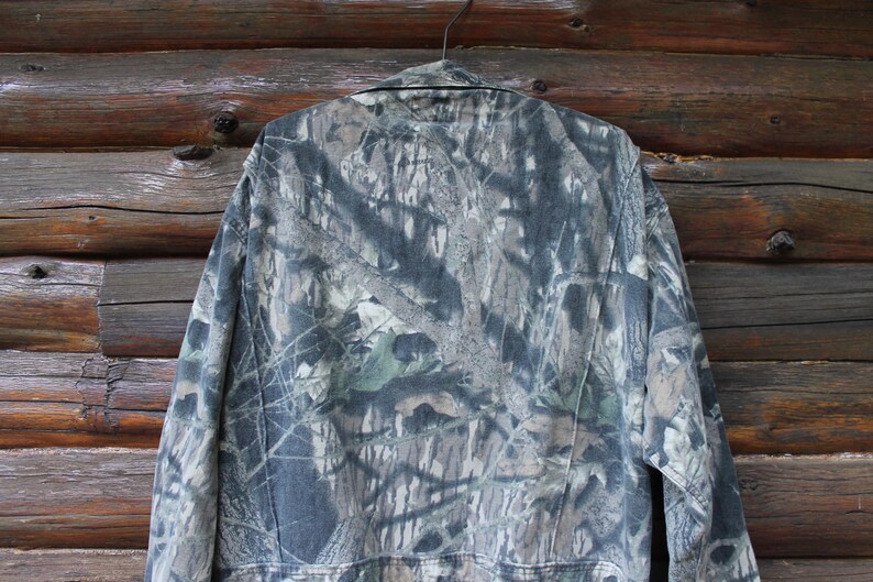 May include: A camouflage jacket hangs on a hanger against a wooden background. The jacket features a pattern of greens, browns, and greys, typical of hunting apparel. The jacket has a collar and buttoned front.