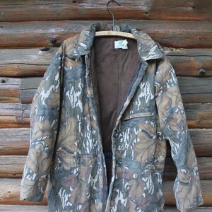 Puede incluir: Una chaqueta de caza con estampado de camuflaje y forro interior marr&oacute;n cuelga de una percha de madera contra una pared de madera r&uacute;stica. La chaqueta tiene cierre de botones, dos bolsillos delanteros y un cuello. El dise&ntilde;o de camuflaje incorpora tonos de marr&oacute;n, verde y blanco.