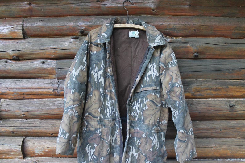Puede incluir: Una chaqueta de caza con estampado de camuflaje y forro interior marr&oacute;n cuelga de una percha de madera contra una pared de madera r&uacute;stica. La chaqueta tiene cierre de botones, dos bolsillos delanteros y un cuello. El dise&ntilde;o de camuflaje incorpora tonos de marr&oacute;n, verde y blanco.