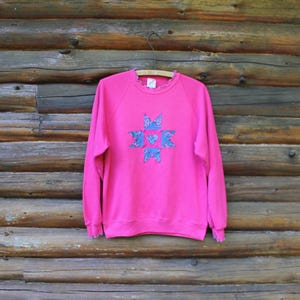 Peut inclure: Un sweat-shirt à manches longues rose vif orné d'une application centrale en forme d'étoile en tissu floral bleu. Le vêtement présente un col rond et des poignets côtelés, tous deux finis par un délicat bord volanté à motif floral bleu.